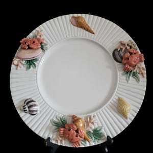 May include: A round white ceramic plate with a ribbed rim featuring three-dimensional decorative elements including orange crabs, various seashells, and light green seaweed accents arranged around the central smooth white surface.