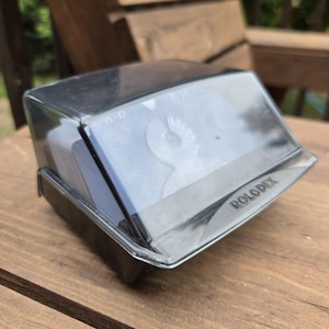 May include: A black Rolodex card file organizer featuring a transparent, hinged plastic cover. The unit contains white index cards with visible A-D lettering. The brand name Rolodex is embossed on the front lower edge of the clear plastic lid.