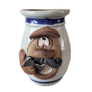 May include: A whimsical ceramic vase with a face design. The vase features a light blue and white body with a blue stripe near the top. The face has a brown hat, mustache, and a textured, tan-colored face with large white eyes.