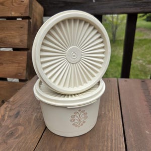 May include: Off-white, vintage Tupperware container with a lid. The lid has a sunburst design. The container has a decorative floral pattern on the side. The container is made of durable plastic and is designed for food storage.