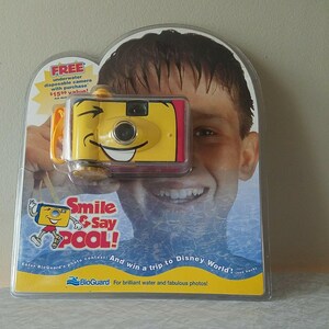 May include: A yellow and red underwater disposable camera with a smiling face. The camera is being held by a boy with wet hair. The text on the packaging reads "Free underwater disposable camera with purchase $15.99 value!" and "Smile & Say POOL! Enter BioGuard's photo contest! And win a trip to Disney World! BioGuard. For brilliant water and fabulous photos!"