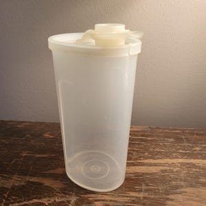 May include: A clear, plastic container with a white lid and a spout. The container is cylindrical with a slightly rounded bottom. The lid has a small, off-white spout and a hinged closure. The container is sitting on a wooden surface.
