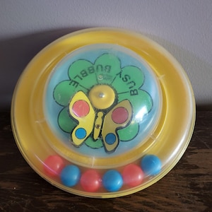 May include: A round yellow plastic toy with a clear dome center. Inside the dome is a green flower graphic with the text BUSY BUBBLE and a yellow butterfly with red and blue wing spots. Six small red and blue balls sit in a circular track.