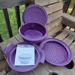 Puede incluir: Un juego de vaporera Tupperware morado que consta de una base, un inserto de colador y una tapa. El juego incluye un manual de instrucciones con el texto Tupperware SmartSteamer Vaporera Inteligente Intelli-Vap y una ilustración.