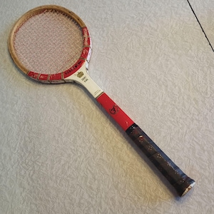 May include: Vintage tennis racket with a wooden frame and a red, white, and black handle. The racket head has a woven string pattern. The handle is wrapped in dark brown grip tape. The racket has the words "Court King" and "Fellmann" printed on it.