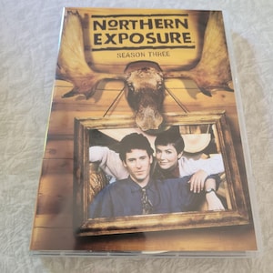 May include: DVD case for the television series "Northern Exposure: Season Three." The cover features a framed photo of two people, with a moose head and the show's logo above. The case is sealed in clear plastic.