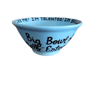 Puede incluir: Un cuenco de cerámica azul claro con letras negras. El cuenco tiene las palabras "Like me! I'm talented! I'm brilliant" alrededor del borde. Las palabras "Big Bowl of Self-Esteem" están escritas en el lateral.