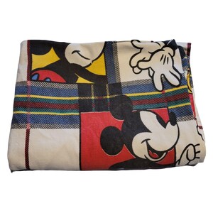 Peut inclure: Couverture pli&eacute;e avec un motif r&eacute;p&eacute;t&eacute; de visages et de mains de Mickey Mouse sur un fond cr&egrave;me. Le motif int&egrave;gre un motif &agrave; carreaux avec des rayures rouges, jaunes, bleues et vertes. La couverture est pli&eacute;e et semble douce.