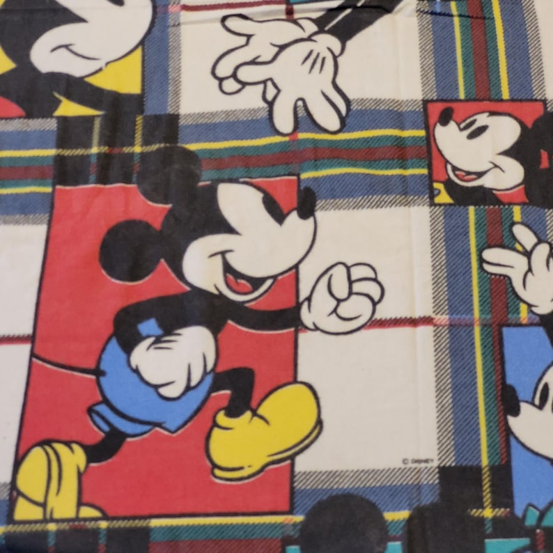 Peut inclure: Tissu avec un motif r&eacute;p&eacute;titif de Mickey Mouse dans diverses poses. Le fond est un motif &agrave; carreaux en blanc, bleu, rouge, jaune et vert. Mickey porte un short bleu et des chaussures jaunes.