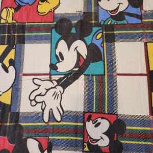 Peut inclure: Un tissu avec un motif r&eacute;p&eacute;titif de Mickey Mouse dans diff&eacute;rentes poses, sur un fond &agrave; carreaux. Les carr&eacute;s ont une base blanche avec des rayures bleues, rouges et jaunes. Le logo Disney est visible en haut &agrave; droite.