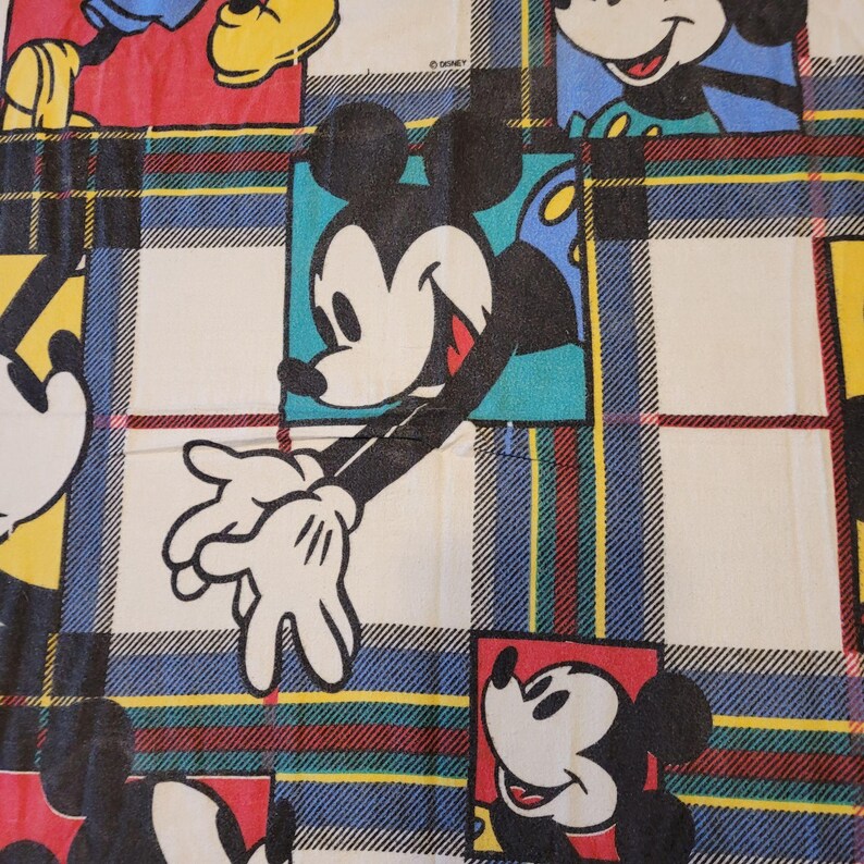 Peut inclure: Un tissu avec un motif r&eacute;p&eacute;titif de Mickey Mouse dans diff&eacute;rentes poses, sur un fond &agrave; carreaux. Les carr&eacute;s ont une base blanche avec des rayures bleues, rouges et jaunes. Le logo Disney est visible en haut &agrave; droite.