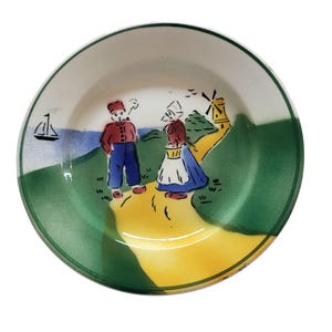 May include: A small, round ceramic plate with a hand-painted Dutch scene. The plate features a green border, a yellow path, and illustrations of a windmill, a sailboat, and two figures in traditional Dutch attire. The plate is white on the inside.