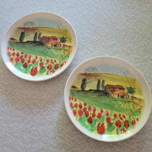 以下が含まれることがあります： 水彩画風の風景画が描かれた2枚の白い丸皿。赤いチューリップ畑、緑の丘、家、糸杉が描かれており、かすかな筆記体の文字と円形の消印スタンプがアクセントになっています。