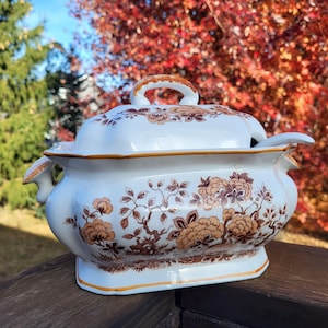 May include: A white ceramic tureen with a matching lid and a side handle. The piece features a brown floral pattern depicting trees and blossoms. It has a rectangular base with rounded corners and a gold-toned trim along the edges and handle.