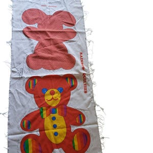 May include: A white fabric panel featuring a printed sewing pattern for a teddy bear. The top section shows a red and white polka dot bear shape, while the bottom displays a finished red bear with a yellow belly, blue buttons, and rainbow accents.