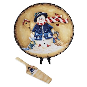 May include: A round, decorative plate featuring a snowman design with a blue coat, hat, and red and white scarf. The plate includes a matching server. The plate is supported by a black stand.