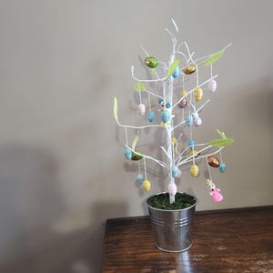 May include: A decorative Easter tree with white branches, adorned with colorful hanging Easter eggs and small figurines. The tree is planted in a silver metal bucket filled with green moss. The tree is placed on a wooden surface.