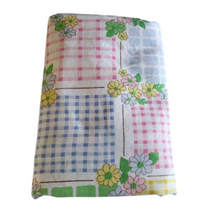 Könnte beinhalten: Gefaltetes Vintage-Blumenbettlaken. Das Laken hat einen weißen Hintergrund mit einem Patchwork-Design aus rosa, blauen und gelben Vichy-Quadraten. Blumencluster in Gelb, Rosa und Weiß sind über das Laken verstreut. Das Laken ist gefaltet.