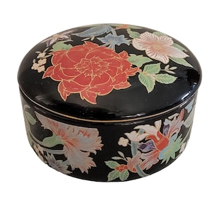 Puede incluir: Una caja redonda de cerámica negra con tapa, adornada con coloridos diseños florales. Las flores son en tonos rojos, rosas y blancos, con hojas verdes y azules. La caja tiene un acabado brillante.