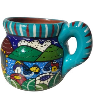Może przedstawiać: Ręcznie malowany ceramiczny kubek o żywym, kolorowym wzorze. Kubek ma turkusowy uchwyt i obrzeże, z namalowaną sceną krajobrazu jeziora, gór i domów. Kubek ma około 10 cm wysokości.
