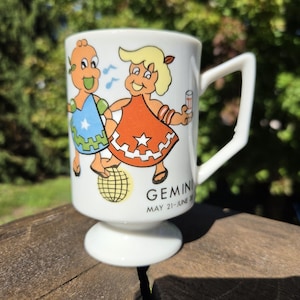 Op de afbeelding: Een witte keramische mok met een voetstuk en een hoekig handvat. De voorzijde toont een kleurrijke afbeelding van twee stripfiguren die dansen bij een gele discobal. De tekst luidt GEMINI MAY 21-JUNE 20, met blauwe muzieknoten ernaast.