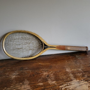 May include: Vintage wooden tennis racket with a light-colored frame and a worn, stringed net. The handle is a darker wood with a leather grip. The racket has black and gold accents near the head. The racket is resting on a dark wooden surface.