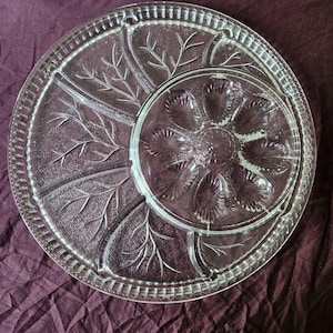 Op de afbeelding: Een serveerschaal van helder glas met een grote ronde schaal met een gestructureerd takkenpatroon en een kleinere, uitneembare ronde inzet in het midden. De inzet heeft een radiaal, schelpachtig ontwerp met acht afzonderlijke holle delen.