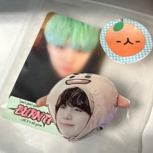 May include: A clear acrylic charm featuring a cartoon character wearing a brown and beige hooded hat. The character has black hair and brown eyes. The charm is on a white background with a sticker that says "yeah yeah yeah BURNT -till it's all gone".