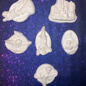May include: Six white plaster figurines, each representing a different fantasy theme. The collection includes a castle, a witch, a figure on a broom, and other characters. The background is a dark blue surface with a glittery effect.