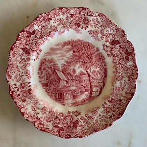 May include: A cream-coloured plate with a scalloped edge and a red floral and landscape design. The centre features a detailed red illustration of a rural scene with a watermill. The rim is adorned with a repeating floral pattern.