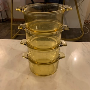 Może przedstawiać: Stos czterech bursztynowych szklanych naczyń do zapiekania z uchwytami. Naczynia są okrągłe i wydają się puste. Są ułożone jeden na drugim na białej marmurowej powierzchni.