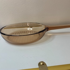 Puede incluir: Una sartén de vidrio color ámbar con un mango largo. La sartén tiene una forma redondeada con un fondo perforado. El mango es una extensión del cuerpo de la sartén. Esta batería de cocina es adecuada para su uso en la estufa.