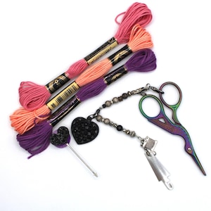 May include: A selection of craft items, including pink, peach, and purple embroidery threads, a black heart-shaped pin, a beaded charm with a black heart, and iridescent scissors. The items are on a white background.