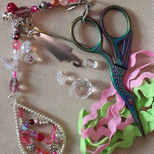 May include: A set of colourful craft supplies including pink and green ribbon, iridescent beads, a silver bead pendant, and a pair of iridescent scissors.