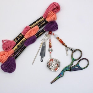 May include: A selection of embroidery tools and materials. There are spools of pink, peach, and purple thread, a decorative charm with beads, and a pair of iridescent scissors. The items are on a white surface, perfect for crafting.