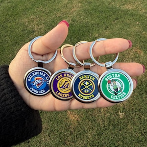 Könnte beinhalten: Eine Sammlung runder Schlüsselanhänger mit NBA-Teamlogos. Die Schlüsselanhänger haben silberne Ringe und farbenfrohe Teamembleme, darunter die Oklahoma City Thunder, Los Angeles Lakers, Denver Nuggets und Boston Celtics.