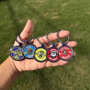 May include: A collection of five round metal keychains featuring college team logos. The keychains have silver rings and colorful designs, including the LSU Tigers, UCLA Bruins, Michigan Wolverines, Georgia Bulldogs, and Ohio State Buckeyes.