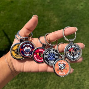 Może przedstawiać: Dłoń prezentuje kilka okrągłych metalowych breloków do kluczy. Każdy brelok ma logo drużyny, w tym Bruins, Oilers, Blackhawks, Kings, Golden Knights i Ducks. Breloki mają srebrne pierścienie.