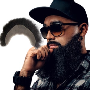 May include: A man with a black beard and a black baseball cap is wearing sunglasses. He is looking down and has his hand on his chin.