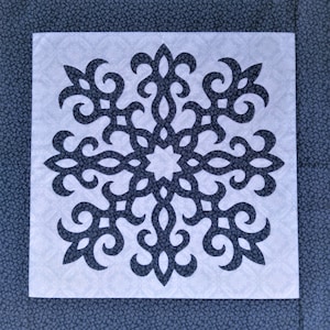 May include: A square quilt block featuring a dark gray, ornate snowflake design on a light gray background. The snowflake is centered within the block, which is bordered by a dark blue fabric with a small floral pattern. The design is symmetrical.