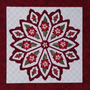 May include: A red and green poinsettia quilt block with a white background. The block is made up of multiple layers of fabric and is quilted with a decorative stitch.