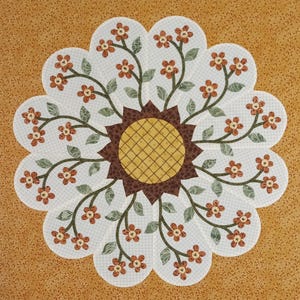 May include: A floral quilt block featuring a central sunflower design surrounded by a scalloped edge. The sunflower has a yellow center with a brown grid pattern, surrounded by brown petals. Delicate stems with small orange flowers and green leaves radiate outwards.