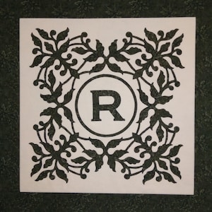 May include: Square fabric art with a white background featuring a black floral design around the edges. A black letter "R" is centered within a circle. The background is a dark green floral pattern.