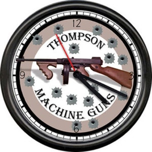 May include: A black framed clock with a beige background featuring a Thompson Machine Guns design. The clock face has a white circle with the words "Thompson Machine Guns" and a silhouette of a machine gun with bullet holes around it.