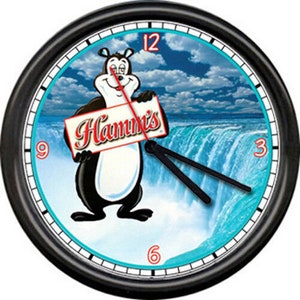 May include: Black wall clock with a white face and black hands. The clock face features a cartoon bear holding a sign that says "Hamm's" and a blue and white image of Niagara Falls.