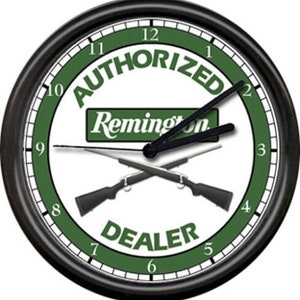 May include: Black and white clock with green accents. The clock face features the text "AUTHORIZED Remington DEALER" with two crossed rifles. The clock hands are black.