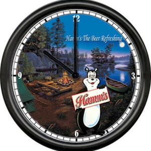 May include: A black wall clock with a picture of a cartoon bear holding a sign that says "Hamm's" in front of a campfire scene. The clock face has a blue background with white numbers and hands. The text "Hamm's The Beer Refreshing" is printed on the clock face.