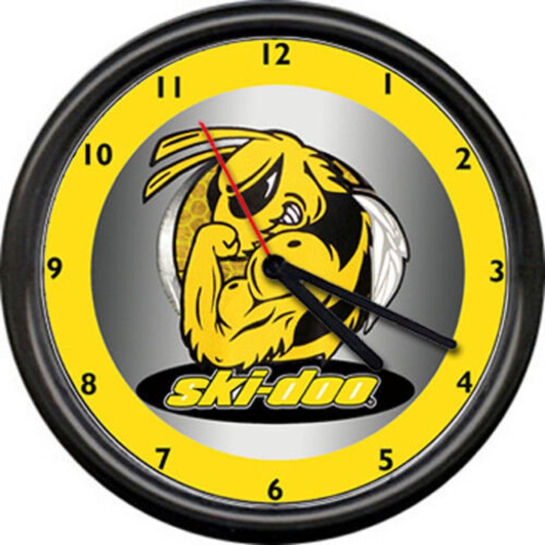 Puede incluir: Un reloj de pared negro y amarillo con un logotipo de Ski-Doo que presenta una abeja de dibujos animados con un brazo musculoso. La esfera del reloj tiene un fondo plateado con n&uacute;meros y manecillas negros.