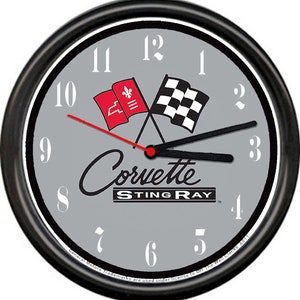 May include: A black and gray wall clock with a silver face. The clock features a red and black Corvette Sting Ray logo with crossed flags. The numbers on the clock face are white.
