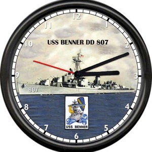 May include: Black clock face with white numbers and a picture of a ship with the text "USS BENNER DD 807" on it. The ship is in the water with a blue sky and white clouds in the background. There is a smaller image of the ship's emblem in the lower right corner with the text "USS BENNER" underneath.